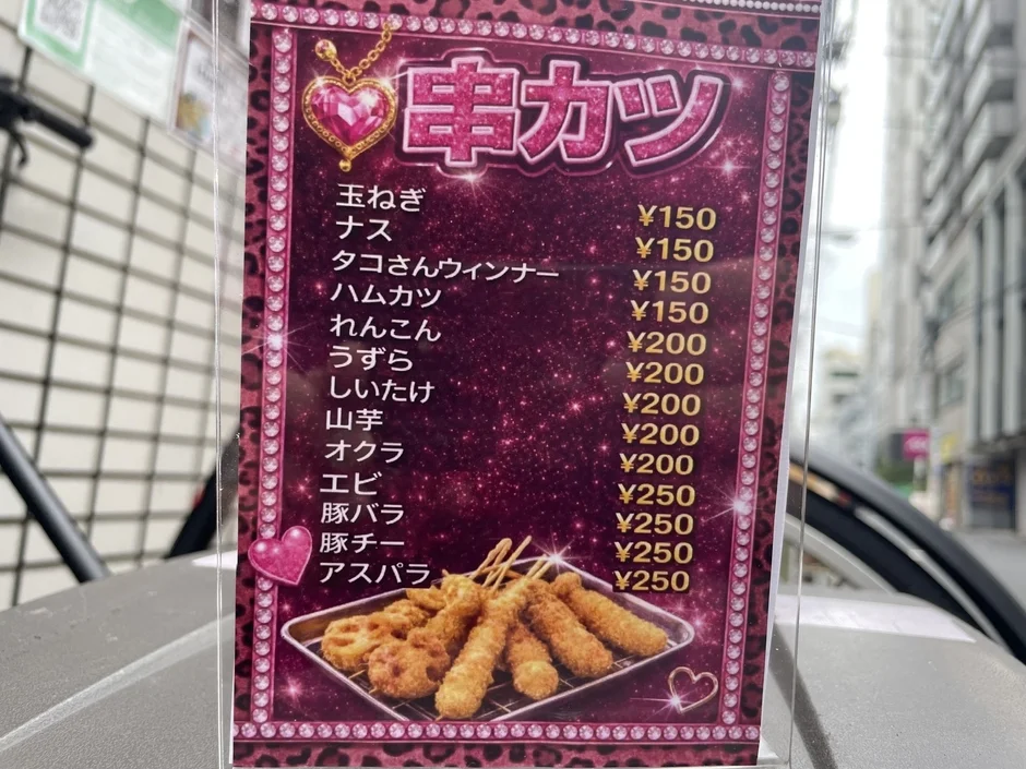 屋外に設置された串カツのメニューです。玉ねぎやナス、エビ、豚バラなど様々な具材が150円から250円で提供されており、ピンクとキラキラしたデザインが特徴です。