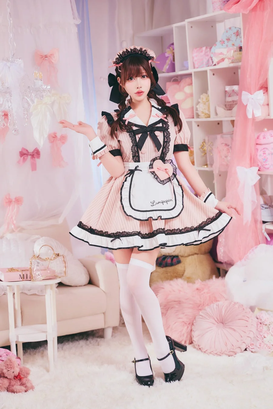 ピンクと黒のメイド服を着た女性が、リボンやぬいぐるみで飾られた可愛らしい部屋でポーズをとっています