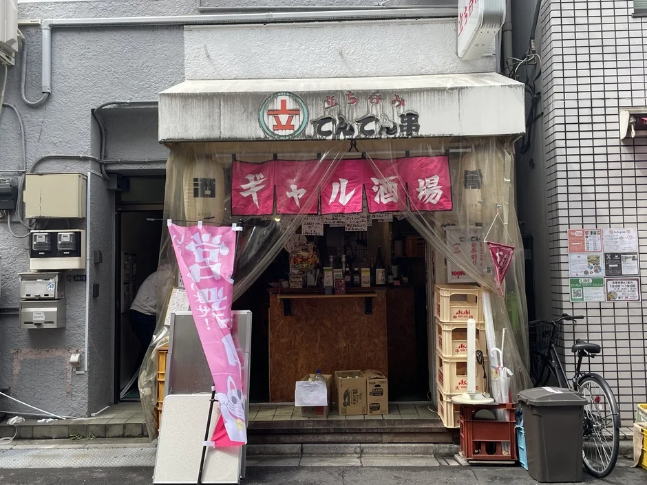 日本の街角にある「立呑み串 ギャル酒場」という立ち飲み屋の店頭。ピンクののぼりやビニールシートが特徴的で、アサヒのケースなどが並び、活気ある雰囲気が感じられる。営業時間は10時から19時で、テイクアウトも可能。