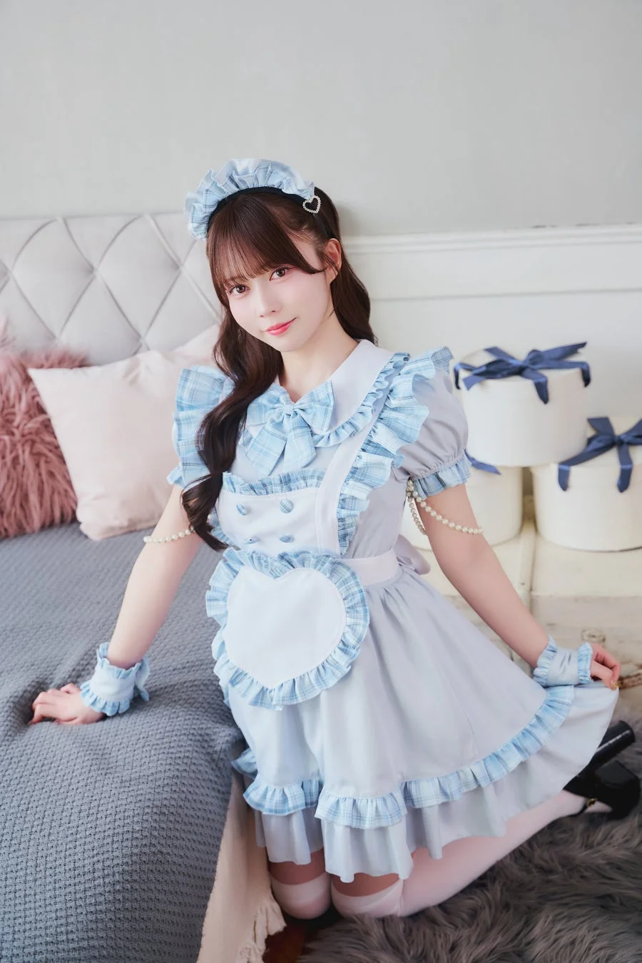 水色と白のフリルが可愛らしいメイド服を着た若い女性が、明るい部屋のベッドの上で膝立ちになり、カメラに笑顔を向けている