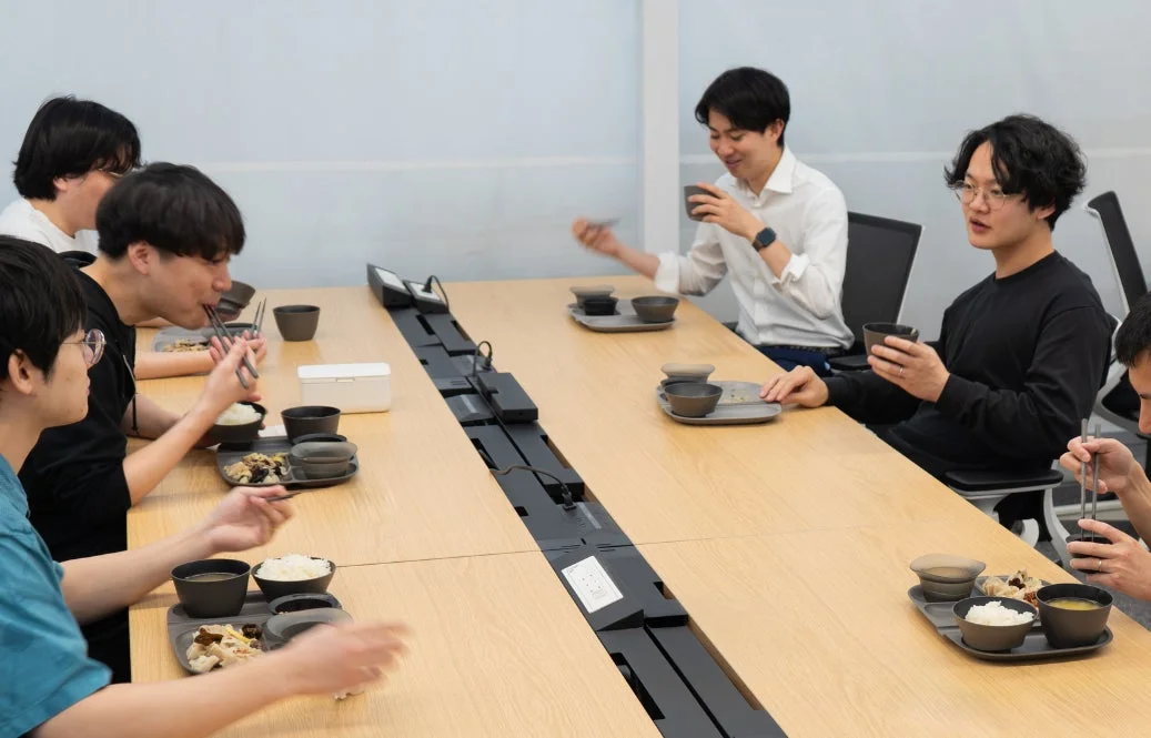 オフィスのような場所で数人の男性が長テーブルを囲んで食事をしている様子