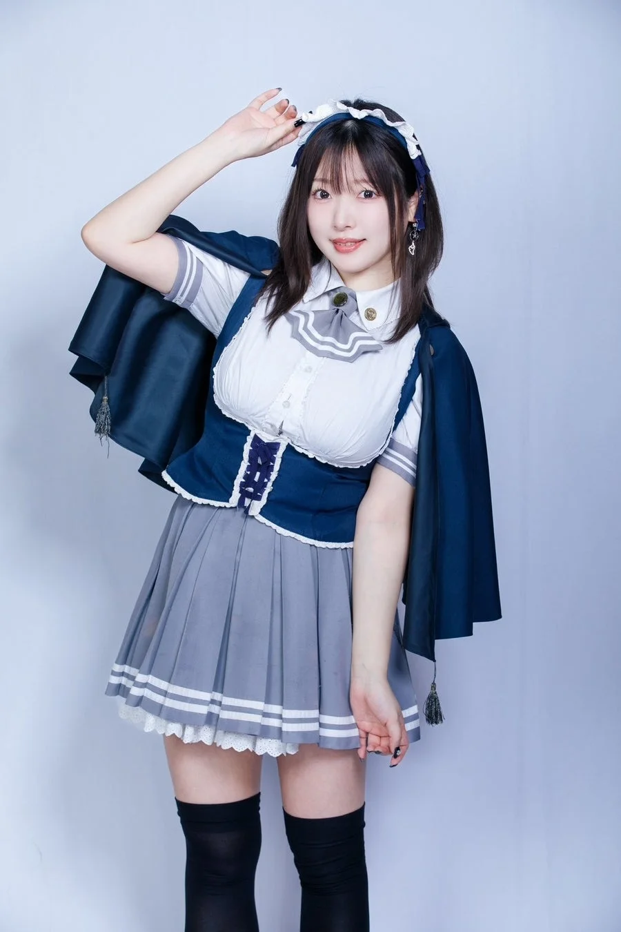 青いケープと制服風の衣装を着用した若い女性が、カメラに向かって笑顔でポーズをとっています。プリーツスカートとニーハイソックスを合わせた可愛らしいスタイルです。