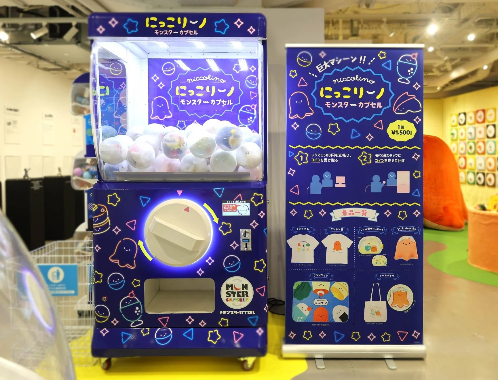 「にっこりーノ モンスターカプセル」と書かれた巨大なガチャガチャマシンと、その景品を紹介する案内板が写っています。1回1500円でTシャツ、ポーチ、ぬいぐるみ、ブランケット、トートバッグなどのオリジナルキャラクターグッズが当たるイベントのようです。