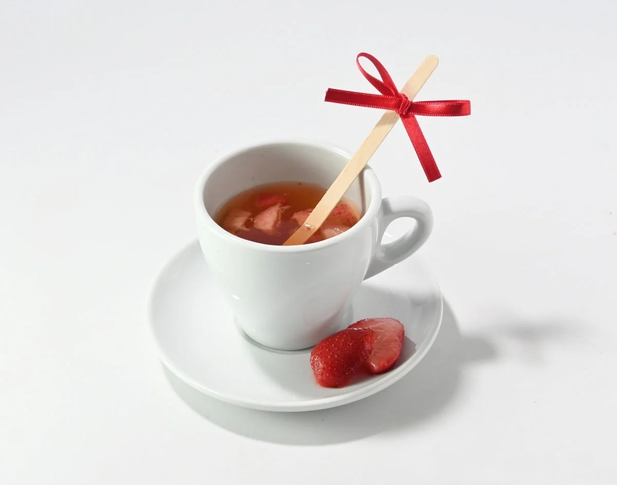白いカップに入ったいちご風味の飲み物で、赤いリボンが付いた木製マドラーが添えられています。ソーサーには新鮮ないちごが添えられており、全体的に清潔感のある白い背景で撮影されています。