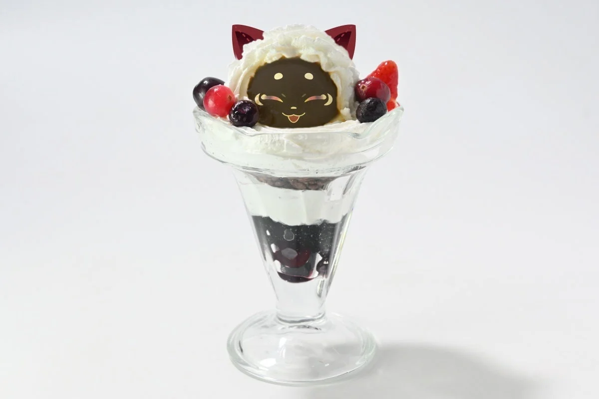 白い背景に置かれた、猫のキャラクターがモチーフの可愛らしいパフェです。ホイップクリームの上にベリーと黒い猫の顔が飾られ、グラスの中には複数の層が見えます。