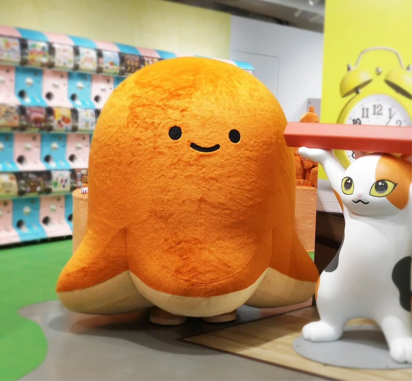 大きなオレンジ色のぬいぐるみが中央にあり、その隣には赤い棒を持つ猫の置物があります。背景にはカラフルなガチャガチャが並び、楽しげなアミューズメント施設の様子を捉えた画像です。