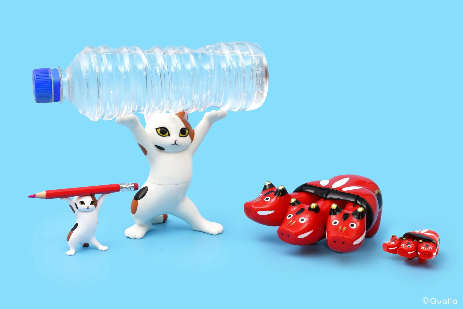 青い背景をバックに、ペットボトルと鉛筆を持ち上げる2匹の猫のフィギュアが並んでいます。その隣には、複数の頭を持つ赤い動物の置物が大小2つ置かれており、ユーモラスな情景を作り出しています。