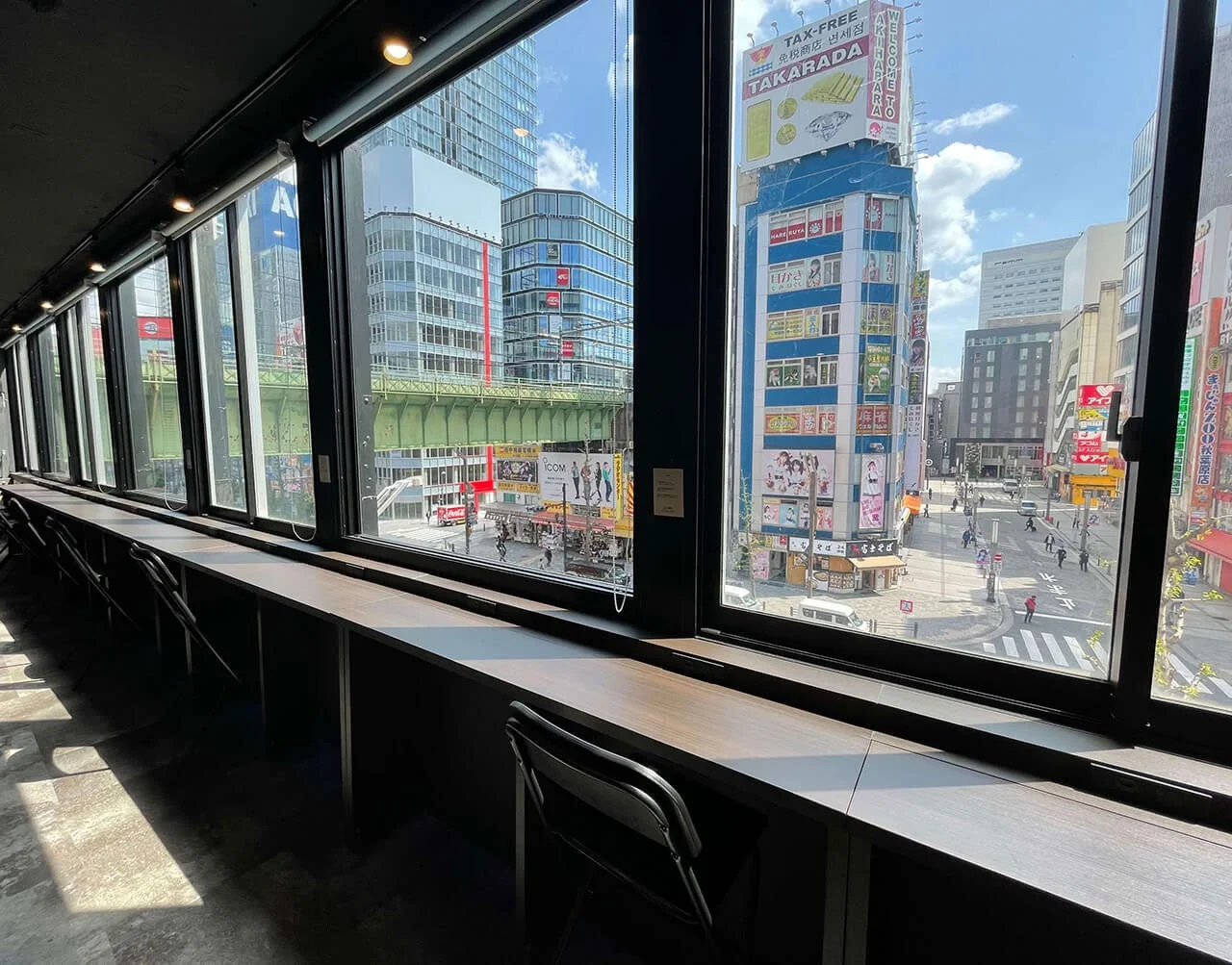 秋葉原の街並みを窓から見下ろす景色。室内にはカウンター席があり、窓の外には多くの商業ビルや鉄道高架、行き交う人々が見える。晴れた日の活気ある都市の風景が広がる。