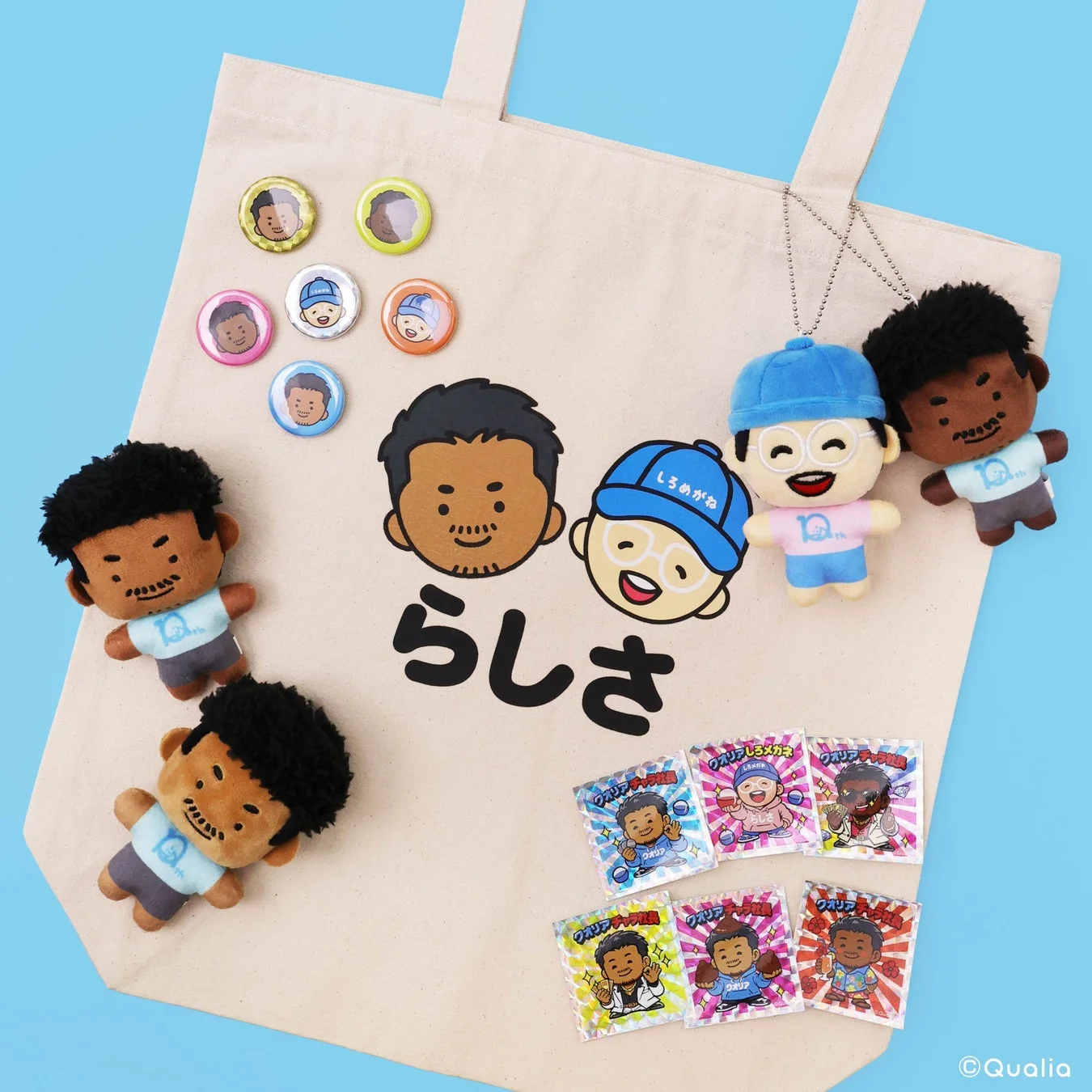 ベージュ色のトートバッグに、可愛らしいキャラクターのぬいぐるみ、缶バッジ、ステッカーが並べられた商品写真。黒髪とメガネの男性キャラクターが特徴で、「らしさ」の文字や「10th」のロゴが見られる。Qualiaブランドのグッズ展開を示すポップな一枚。