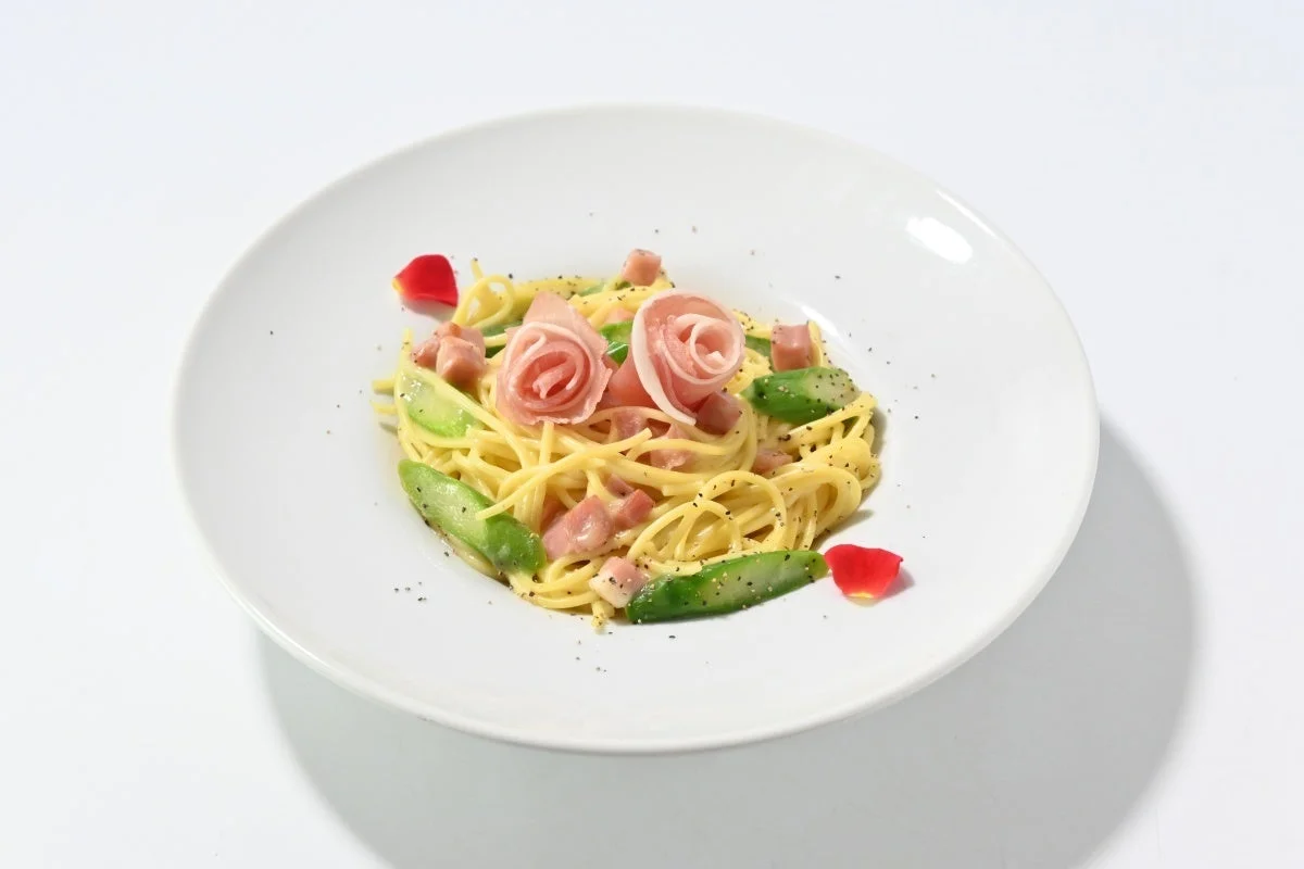 生ハムとアスパラガスを添えたクリームパスタ。生ハムはバラの花のように飾り付けられ、赤いバラの花びらが散らされており、見た目も華やかな一皿です。