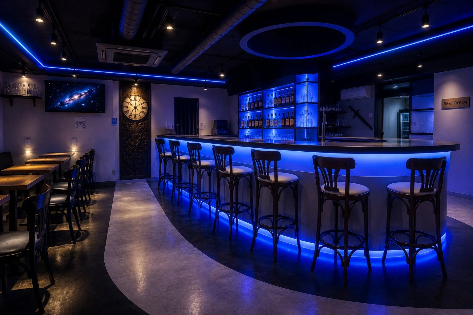 青いLED照明が印象的な、モダンでスタイリッシュなバーの店内です。長いカウンターバーにはボトルが並び、その周りにハイスツールが配置されています。左手にはテーブル席もあり、壁には宇宙の画像が映るテレビと大きな時計が見えます。