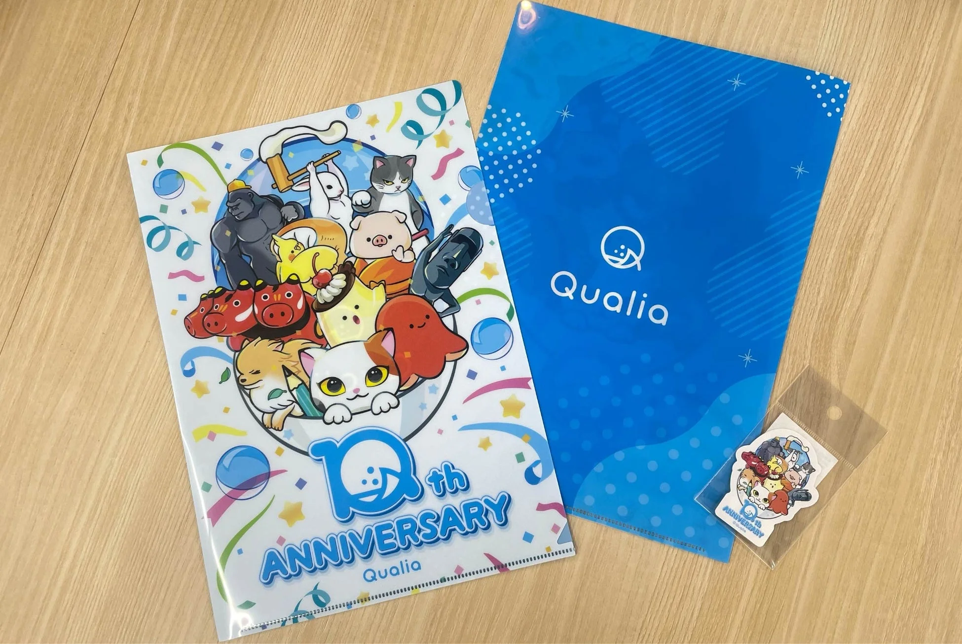 Qualiaの10周年を記念したクリアファイルとステッカーが木製のテーブルに置かれています。クリアファイルには、様々な動物やキャラクターが賑やかに描かれたデザインと、Qualiaのロゴが入ったシンプルな青いデザインの2種類があります。