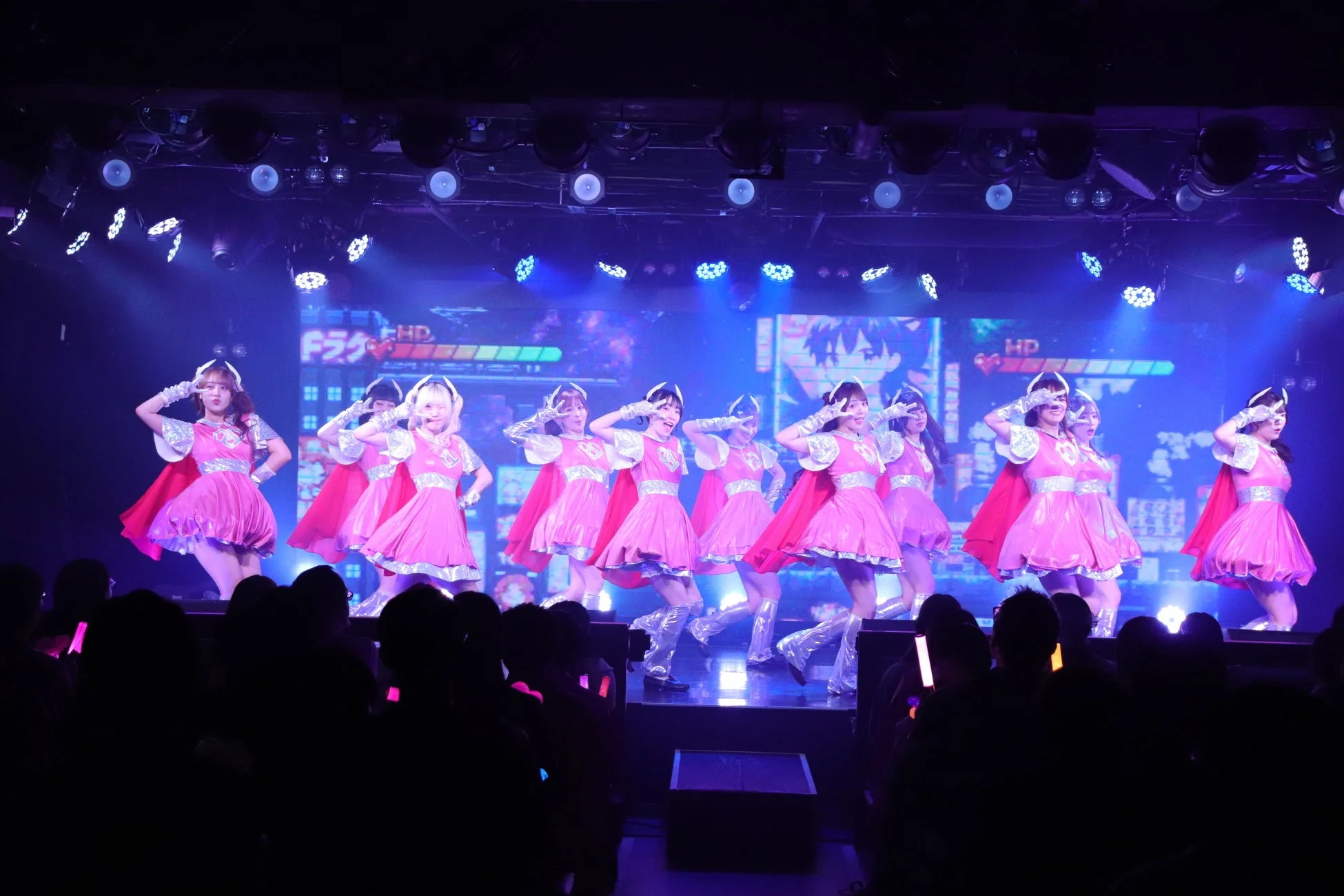 ピンクのスーパーヒーロー風衣装をまとった女性アイドルグループがステージでパフォーマンス中