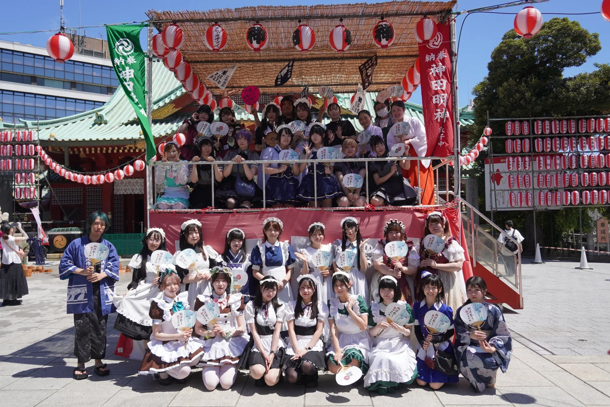 神田明神を背景に、様々なメイド服を着た大勢の女性たちが集合写真を撮っています。提灯や幟で飾られたお祭りムードの中、皆うちわを持ち、笑顔でポーズをとっています。メイド喫茶の周年イベントのようです。