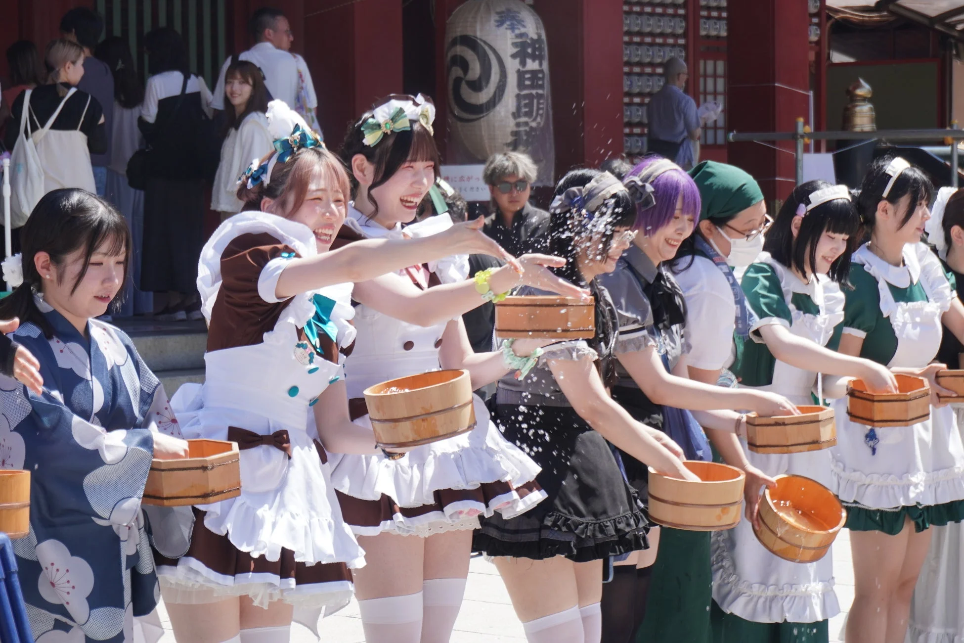 メイド服や浴衣を着た若い女性たちが、屋外のイベントで木製の升から何かを撒き、楽しんでいる様子。背景には「奉納 神田」と書かれた提灯が見え、神田祭に関連する行事のようだ。