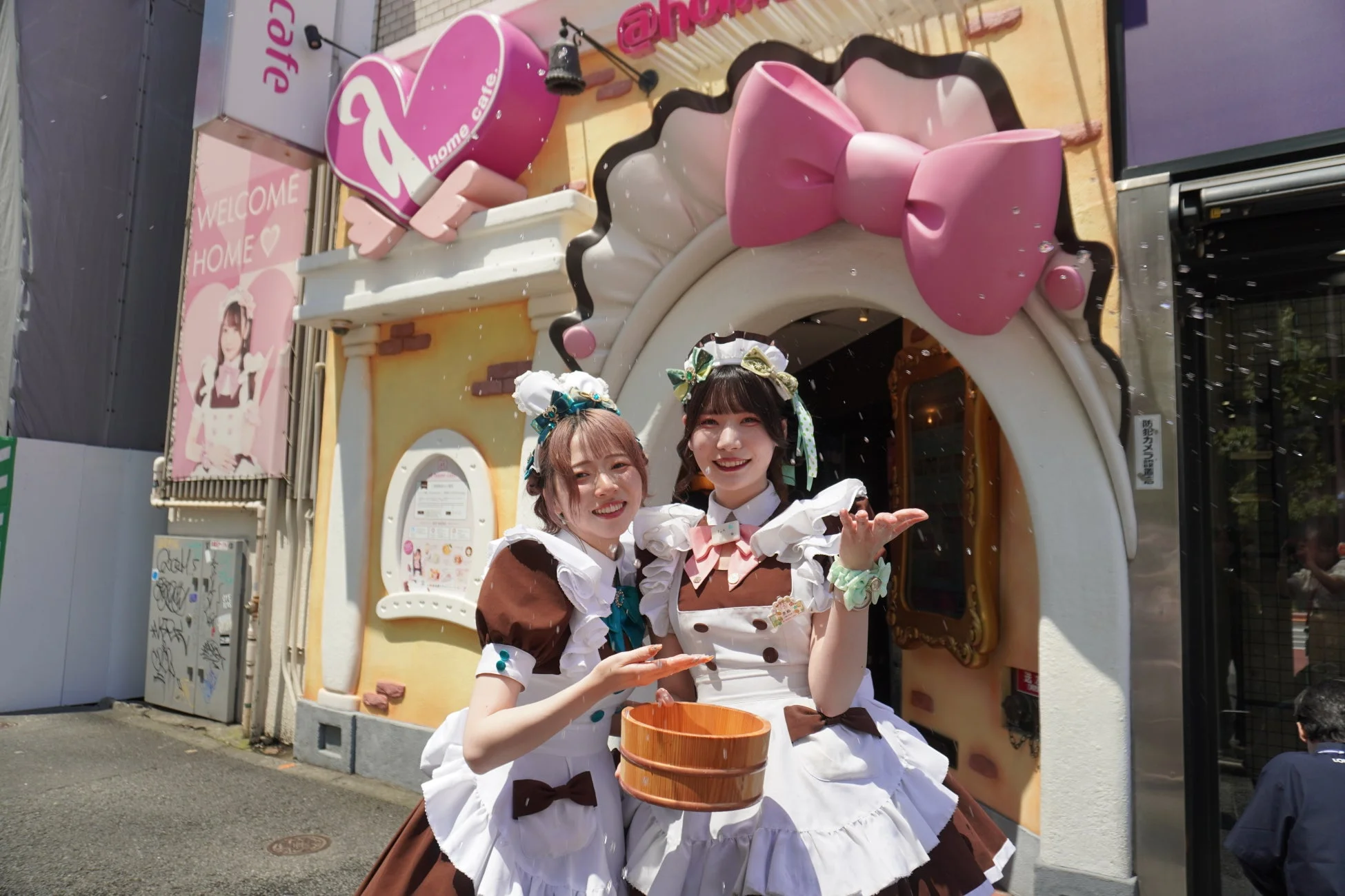 メイド服を着た2人の女性が、大きなピンクのリボンが飾られたメイドカフェの入り口前で笑顔でポーズを取っています。木製の桶を持ち、周囲にはシャボン玉のようなものが舞い、賑やかで可愛らしい雰囲気が伝わります。